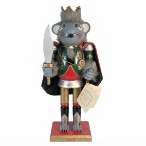 May include: A wooden nutcracker figurine depicting a mouse king wearing a gold crown, a green and red jacket with gold floral details, and a black cape. The mouse holds a silver sword and stands on a red and gold base with the year 2010 printed on it.