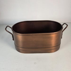 May include: An oval-shaped metal basin with a brushed copper finish. The container features two side handles attached to the exterior and a horizontal embossed line running along the lower portion of the body, providing a simple, functional design.