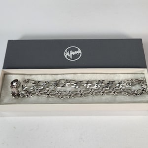 May include: A silver-toned necklace and matching earrings set displayed in an open gray box with a white interior. The necklace features a chain link design. The box has a logo on the lid.