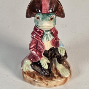 May include: A ceramic figurine of a green frog dressed in a maroon frock coat, a white cravat, and a brown bicorne hat. The frog is seated on a brown base, with one hand resting on a knee, featuring a glossy finish and detailed painted features.