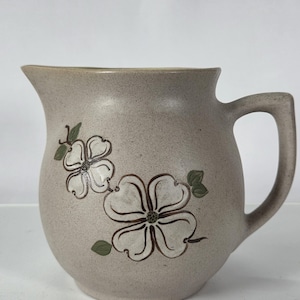 May include: A beige ceramic pitcher with a rounded body and a handle. The pitcher is decorated with two white dogwood flower designs with brown outlines and green leaves. The pitcher has a speckled texture and a wide spout.