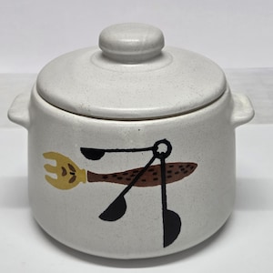 May include: A speckled, off-white ceramic jar with a lid and two handles. The jar features a whimsical design of a yellow fork, black spoons, and a brown fish-shaped element. The lid has a small, rounded knob.