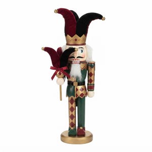 May include: A nutcracker figurine dressed as a jester in a velvet burgundy and black hat with gold bells. The figure wears a green and gold diamond-patterned outfit, holds a matching jester staff, and features a white beard and mustache.