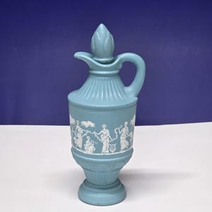May include: A light blue ceramic pitcher with a handle and a decorative stopper. The pitcher features a white relief design of figures in classical attire. The pitcher is approximately 7 inches tall.