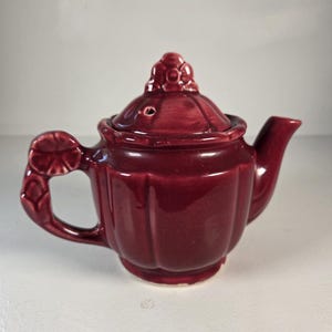 May include: A deep burgundy ceramic teapot featuring a fluted body, a matching lid with a floral-shaped finial, and a handle adorned with a molded flower detail. The teapot has a glossy finish and a small steam vent on the lid.