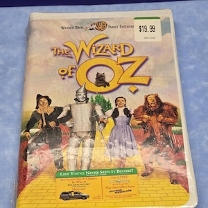 May include: A VHS tape of "The Wizard of Oz" movie. The cover features the film's title in gold lettering, with illustrations of the Scarecrow, Tin Man, Dorothy, and the Cowardly Lion. The tape is sealed in plastic.