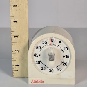 May include: A white Sunbeam kitchen timer with a circular dial marked from 0 to 55 minutes, featuring a clear plastic knob. A wooden ruler is positioned vertically next to the timer, showing the device stands approximately 3.5 inches tall.