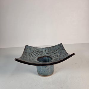 May include: A ceramic candle holder with a square, flared top featuring a dark blue and black textured lace-like pattern. The base is a cylindrical, speckled blue pedestal. The edges of the square top are dark, contrasting with the blue center.