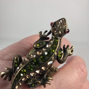 May include: A lizard-shaped ring crafted from antique bronze-toned metal, featuring a textured body encrusted with small, sparkling green and clear rhinestones. The lizard has two small, bright red rhinestones for eyes and a hinged, adjustable band.