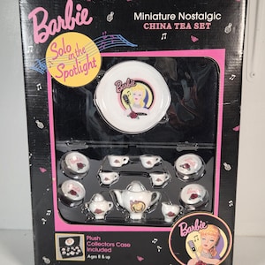 May include: A Barbie Miniature Nostalgic China Tea Set in a black box with pink accents. The set includes a teapot, cups, saucers, and a creamer, all featuring a graphic of a blonde figure with a microphone. The box text reads Solo in the Spotlight.