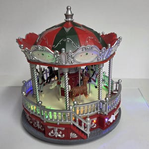 May include: A miniature carousel with a red and dark green roof, silver accents, and a cream-colored base. The structure features multiple animal figures, including a brown deer, and is illuminated by small, colorful LED lights along the platform.