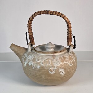 May include: A round ceramic teapot with a beige and white mottled glaze, featuring a textured surface and a removable lid. The teapot has a curved spout and a tall, arched handle wrapped in brown woven rattan, secured with black cord accents.