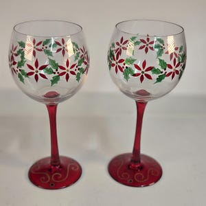 May include: Two clear glass wine glasses featuring a hand-painted holiday motif of red poinsettia flowers and green holly leaves around the bowl. The stems are solid red, and the circular bases feature decorative gold swirl accents.