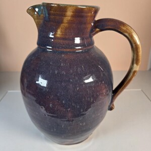 May include: A handcrafted ceramic pitcher featuring a rounded body and a curved handle. The glaze displays a deep purple and dark brown color palette with subtle variations in tone and a glossy finish, including a distinct pouring spout.