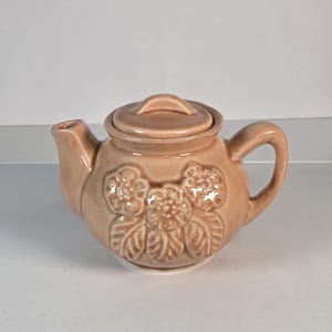 May include: A light peach-colored ceramic teapot featuring a raised, embossed floral design on the front. The teapot has a rounded body, a curved handle, a short spout, and a matching removable lid with a small central knob handle.
