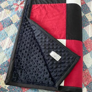 May include: A patchwork quilt in a colour block design of black, red, and white. The quilt has a soft, black, textured backing and a black border. It is folded, showing the plush backing and a small label.