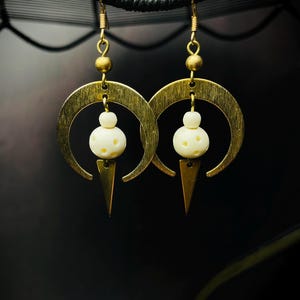 Peut inclure: Une paire de boucles d'oreilles en or avec un motif de croissant de lune et un pendentif en perle blanche. Les boucles d'oreilles ont une pointe pointue en bas.