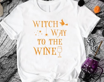 May include: White t-shirt with orange text that reads "Witch Way to the Wine" with a witch on a broom, stars, a spider, and a wine glass. The shirt is styled with Halloween decorations.