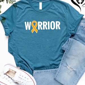 May include: Teal-colored t-shirt with the word "WARRIOR" in white block letters and a yellow ribbon graphic. The shirt is paired with light blue denim jeans and white sneakers with red and blue accents.