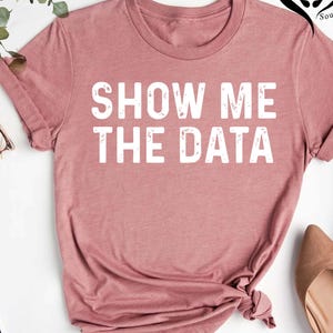 May include: A dusty rose-colored t-shirt with the phrase "SHOW ME THE DATA" printed in bold white capital letters. The shirt has short sleeves and a crew neck. The shirt is knotted at the bottom.