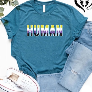 May include: Teal t-shirt with the word "HUMAN" in large, bold letters. The letters are colored in the non-binary pride flag colors: yellow, purple, black, and white. The shirt is paired with light-wash denim jeans and white sneakers.