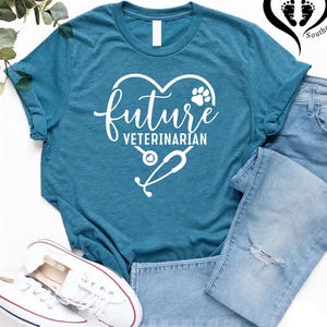 May include: Teal t-shirt with the words "future veterinarian" in a heart-shaped design, including a paw print and stethoscope. The shirt is paired with light blue jeans and white sneakers.