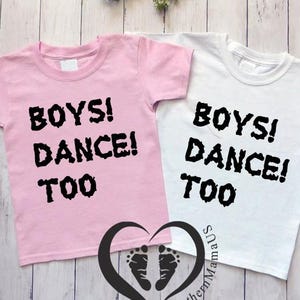 May include: Two short-sleeved t-shirts, one pink and one white, with the text "BOYS! DANCE! TOO" in black. A plush unicorn toy and a small, colorful toy are also visible.