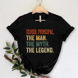 May include: A black short-sleeved t-shirt featuring the text SCHOOL PRINCIPAL. THE MAN. THE MYTH. THE LEGEND. in a distressed, multi-colored font with orange, yellow, and teal lettering centered on the chest. The shirt hangs on a wooden hanger.