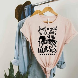 May include: A light pink t-shirt with the text "Just a girl who loves horses" and a black horse graphic. The horse graphic is filled with floral patterns. The shirt is hanging on a wooden hanger with other clothing.