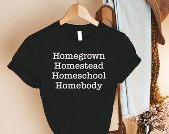 May include: Black t-shirt with the words "Homegrown, Homestead, Homeschool, Homebody" in white block letters. The shirt is hanging on a wooden hanger. A hat and scarf are visible in the background.
