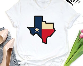 May include: White t-shirt featuring a graphic of the Texas state flag. The design includes a blue field with a white star, a cream-colored section, and a red section, all outlined in black.