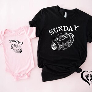 Puede incluir: Una camiseta negra con la palabra "SUNDAY" y un gr&aacute;fico de f&uacute;tbol americano, junto a un body rosa para beb&eacute; con la palabra "FUNDAY" y un gr&aacute;fico de f&uacute;tbol americano a juego. Fondo rosa claro.