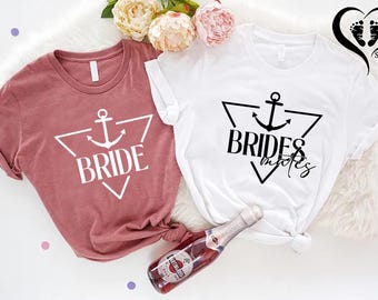 May include: Two t-shirts, one mauve and one white, with anchor designs inside a triangle. The mauve shirt says "BRIDE" and the white shirt says "BRIDESMAIDS". A bottle of sparkling wine is in the foreground.