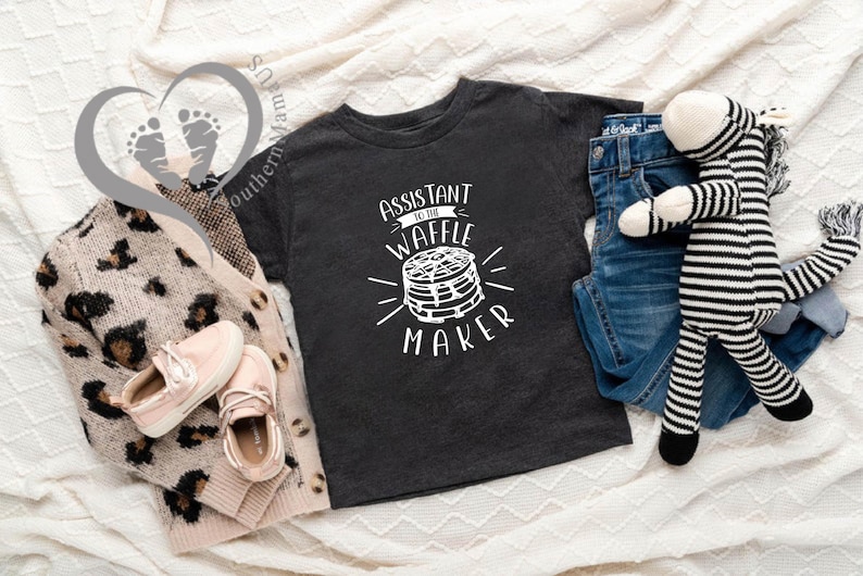 Puede incluir: Una composici&oacute;n plana con una camiseta gris oscuro con el texto "Assistant to the Waffle Maker", vaqueros azules, un peluche de cebra, una rebeca con estampado de leopardo y zapatos rosas. Los art&iacute;culos est&aacute;n dispuestos sobre una superficie blanca texturizada.