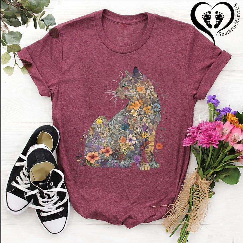 May include: Burgundy t-shirt featuring a floral cat design. The cat is composed of various colorful flowers. Black and white sneakers are on the left. A bouquet of pink and yellow flowers is on the right.