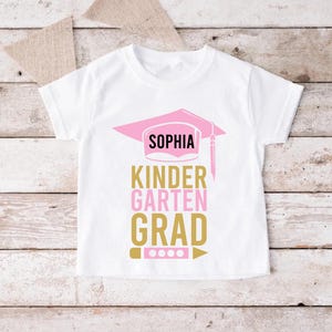 May include: White t-shirt with a pink graduation cap design and the name "Sophia". The text below reads "Kindergarten Grad" in gold and pink. A pencil and dots are at the bottom of the design.