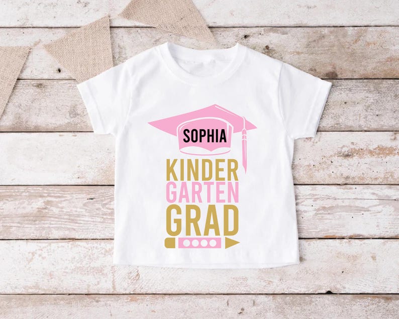 May include: White t-shirt with a pink graduation cap design and the name "Sophia". The text below reads "Kindergarten Grad" in gold and pink. A pencil and dots are at the bottom of the design.