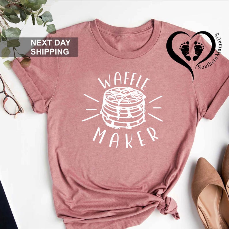 Puede incluir: Camiseta rosa polvoriento con un gr&aacute;fico blanco de una pila de gofres y las palabras "WAFFLE MAKER". La camiseta tiene mangas cortas y cuello redondo. La camiseta est&aacute; anudada en la parte inferior.