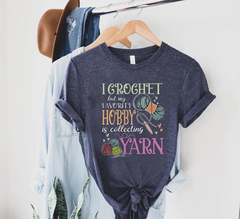 May include: A navy blue t-shirt with the text 'I crochet but my favorite hobby is collecting yarn' in colorful text with a graphic of yarn and knitting needles.