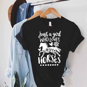 May include: Black t-shirt with the text "Just a girl who loves horses" in white script. A white horse silhouette with floral details is featured in the design. The shirt is hanging on a wooden hanger.
