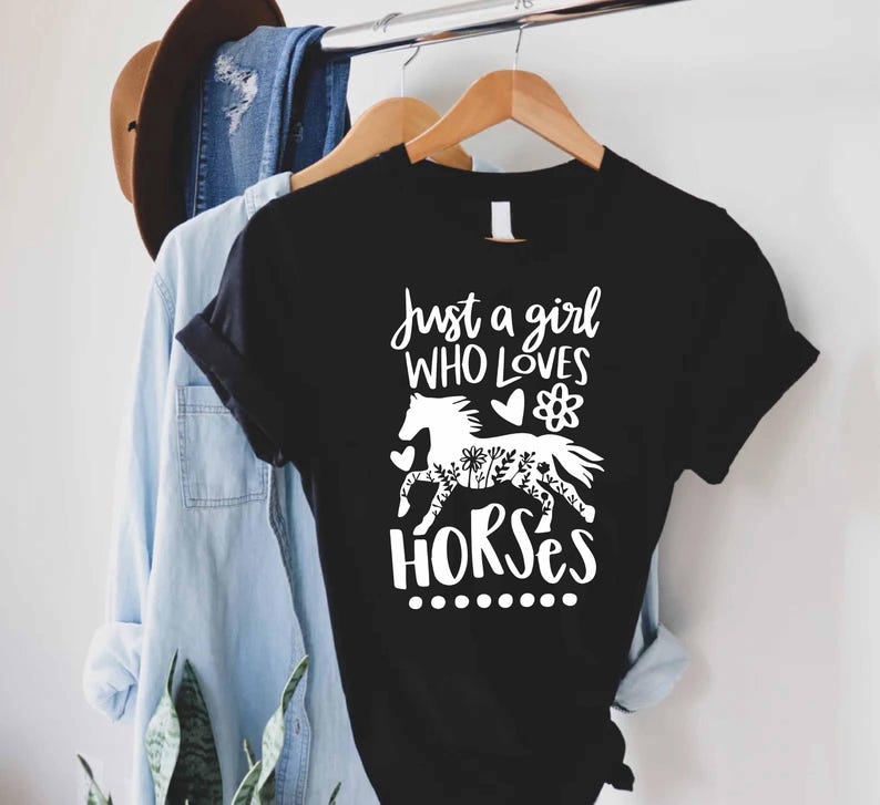 May include: Black t-shirt with the text "Just a girl who loves horses" in white script. A white horse silhouette with floral details is featured in the design. The shirt is hanging on a wooden hanger.