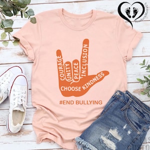 May include: A light pink t-shirt with a graphic of a hand in a sign language gesture. The orange graphic includes the words "Courage, Unity, Peace, Inclusion, Kindness" and "Choose Kindness." The text "#END BULLYING" is below the hand.