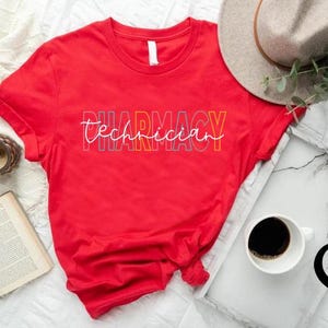 K&ouml;nnte beinhalten: Ein rotes T-Shirt mit dem Text "Pharmacy Technician" in einem farbenfrohen, mehrschichtigen Design. Das Shirt wird mit Hut, Kaffee und anderem Zubeh&ouml;r pr&auml;sentiert, was auf einen Lifestyle- oder Geschenkartikel hindeutet.