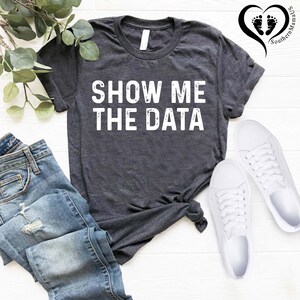 May include: Dark gray t-shirt with the text "SHOW ME THE DATA" in white block letters. The shirt is paired with blue jeans and white sneakers. A sprig of greenery and a logo are also visible.