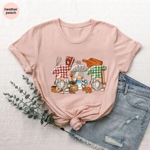 May include: A light pink t-shirt featuring a graphic of three gnomes dressed as bakers, each with a different checkered hat. The gnomes are holding baking tools and a croissant. The text "heather peach" is in the upper left corner.