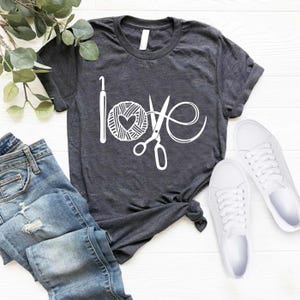 May include: Dark gray t-shirt with the word "love" spelled out with a crochet hook, yarn ball with a heart, and scissors. The shirt is paired with blue jeans and white sneakers, creating a casual, everyday look.
