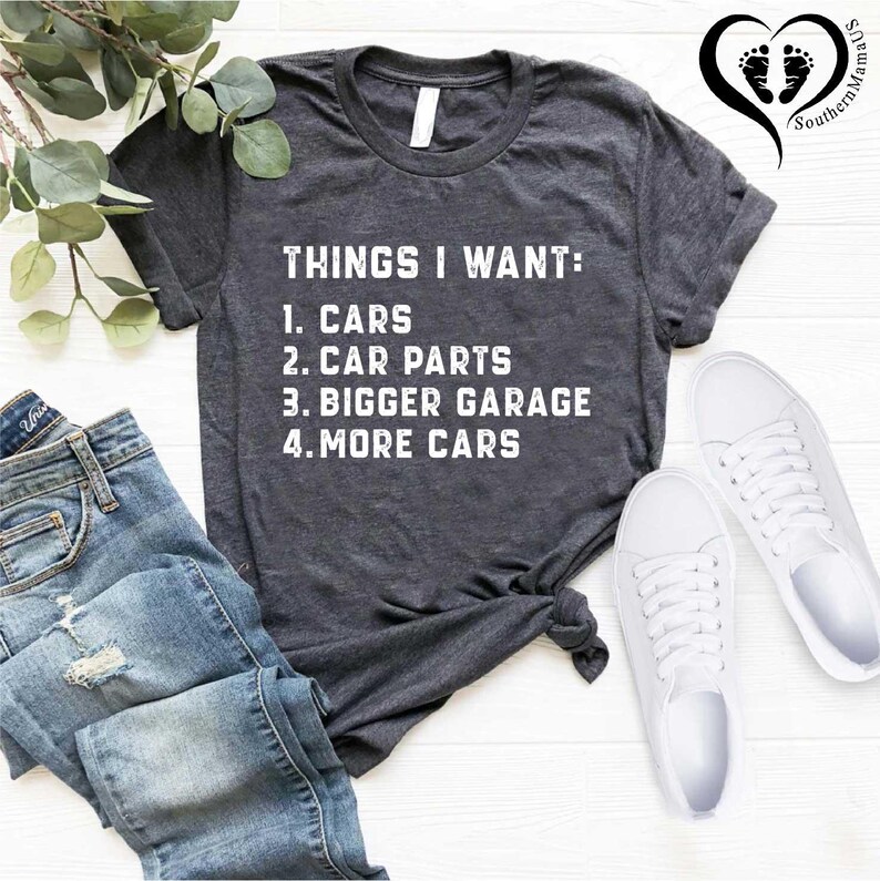 Puede incluir: Una camiseta gris oscuro con texto blanco que dice "THINGS I WANT: 1. CARS 2. CAR PARTS 3. BIGGER GARAGE 4. MORE CARS". La camiseta est&aacute; colocada sobre una superficie blanca con un par de zapatillas blancas y un par de pantalones vaqueros azules.
