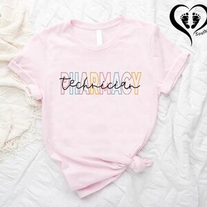 K&ouml;nnte beinhalten: Hellrosa T-Shirt mit dem Aufdruck "Pharmacy Technician" in bunten Buchstaben. Das Wort "Pharmacy" ist in verschiedenen Farben geschrieben. Das T-Shirt ist am unteren Ende geknotet.