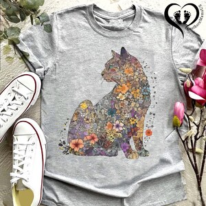 May include: A heather gray t-shirt featuring a seated cat design filled with colorful flowers. The cat's silhouette is detailed with various floral patterns in shades of orange, purple, and yellow. White sneakers are visible on the left side of the shirt.