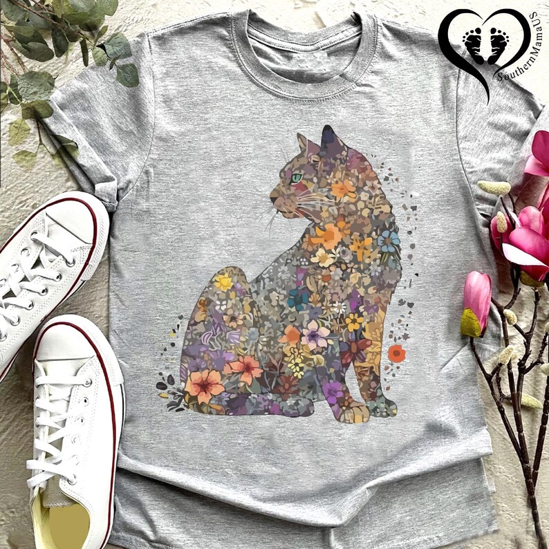 May include: A heather gray t-shirt featuring a seated cat design filled with colorful flowers. The cat's silhouette is detailed with various floral patterns in shades of orange, purple, and yellow. White sneakers are visible on the left side of the shirt.